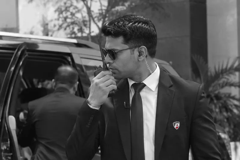 A close protection officer in a dark suit speaking into a handheld radio beside an open car door